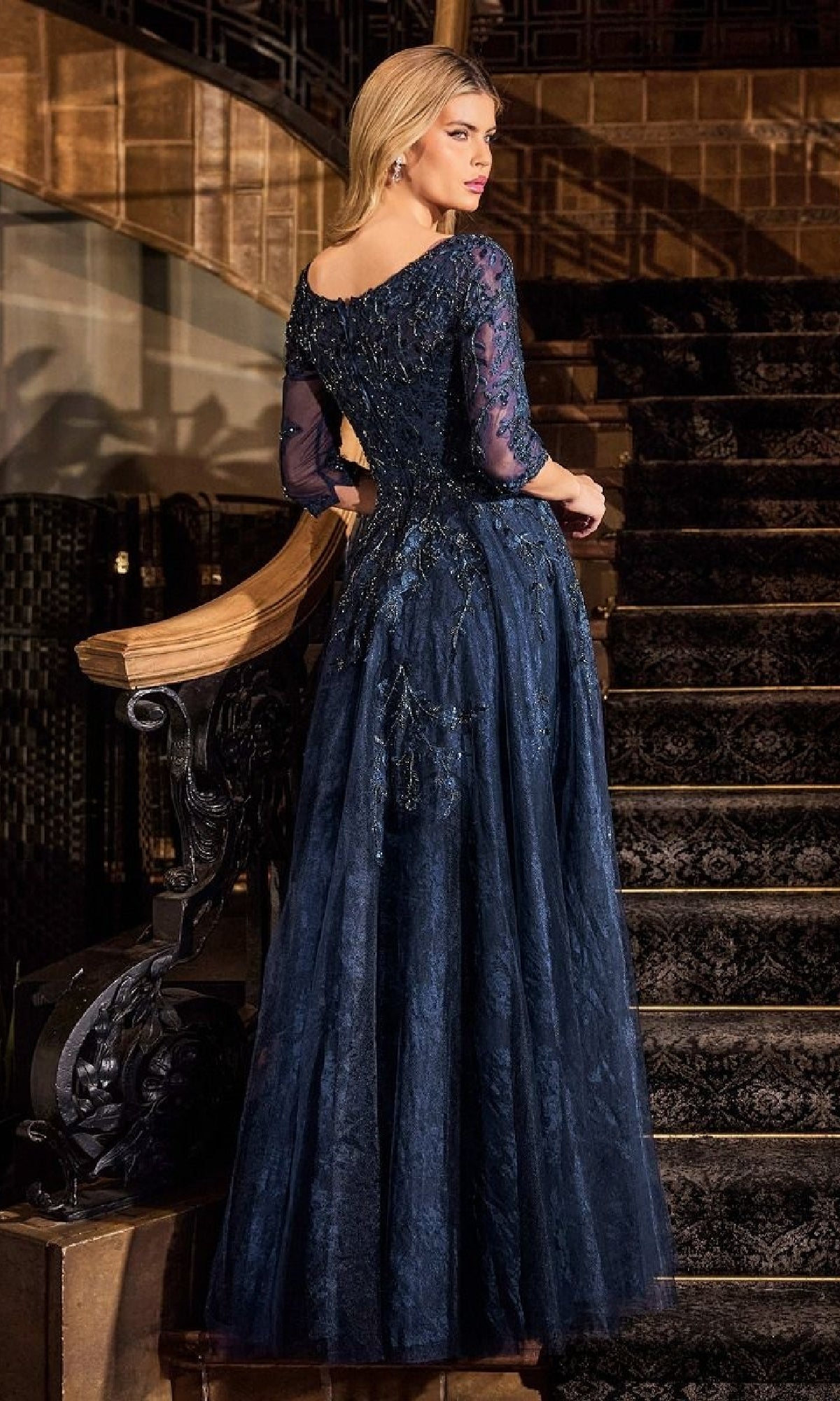 A woman stands on a staircase wearing the Ladivine CK2401, a long sleeve blue lace formal gown, floor-length with sheer sleeves and intricate bead detailing, facing away but glancing over her shoulder.